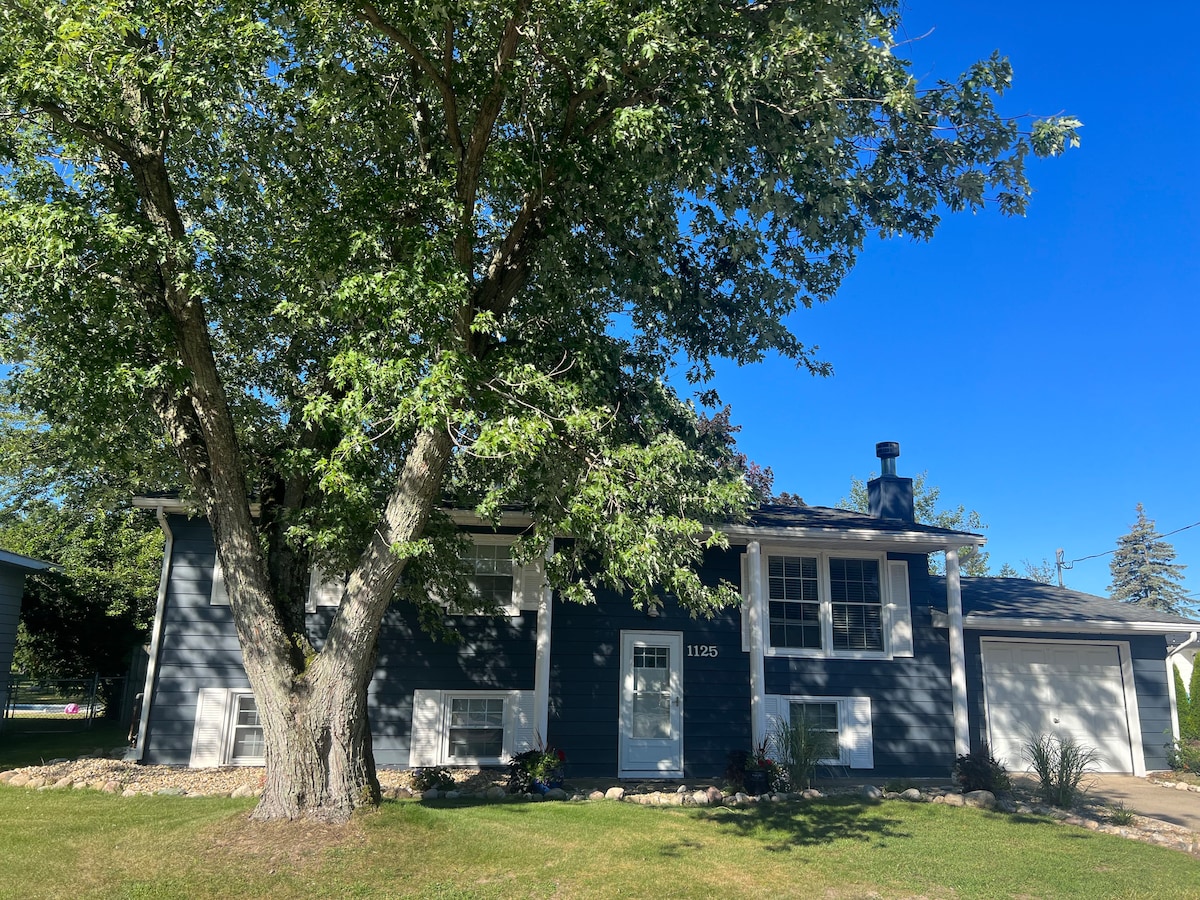 The exterior of a two-story beach house is framed by a large tree, casting shade on the front yard. A welcoming entrance is centered, flanked by a few shrubs. The house features a classic design with blue siding and a chimney visible on the right side.