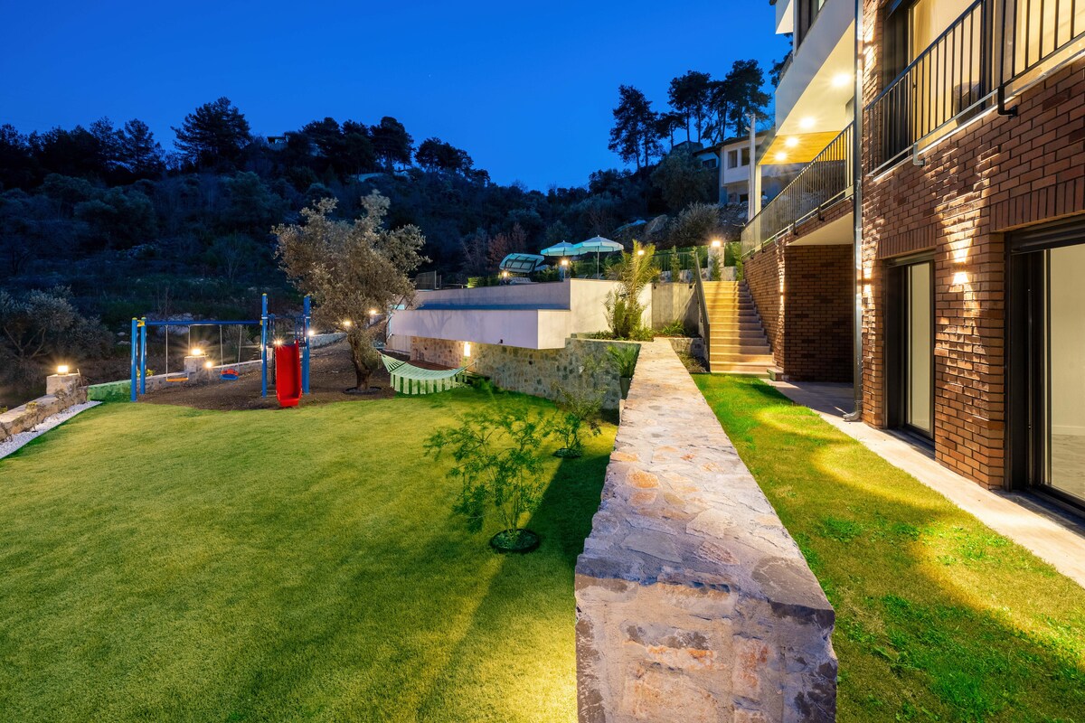 The outdoor area is highlighted by a spacious lawn bordered by stone pathways, leading to a children's play area with swings. Soft lighting enhances the landscape, while the backdrop features trees and illuminated terraces visible from the property.