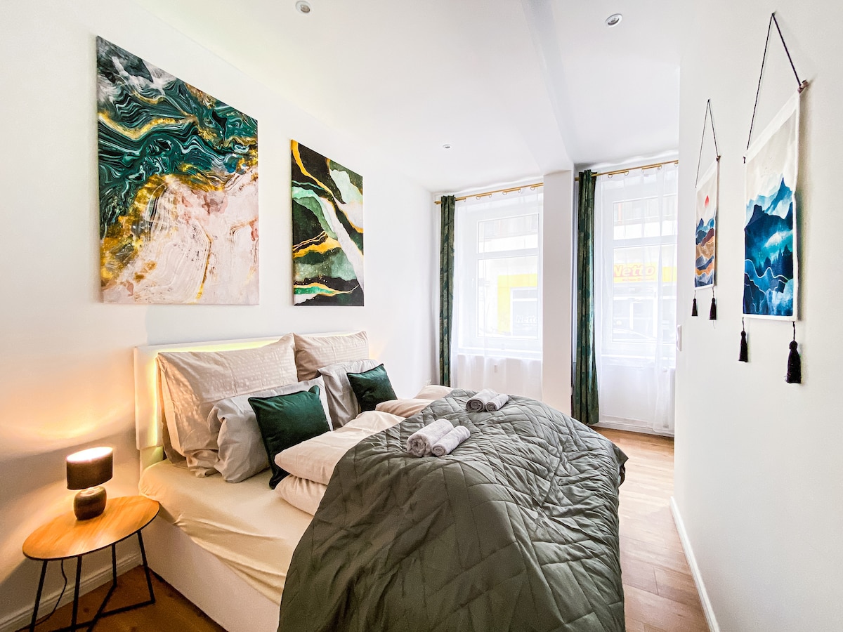 A welcoming bedroom features a spacious bed with soft pillows and a dark green quilt. The walls are adorned with abstract art pieces in shades of green and pink. Natural light enters through large windows, complemented by elegant curtains, creating a calm ambiance.