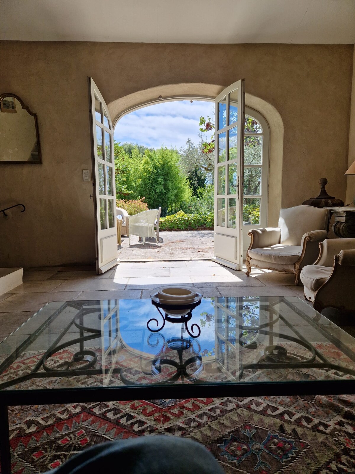 Natural light fills the inviting living space, where an elegantly framed doorway opens onto a serene outdoor area. A glass table is centered on a patterned rug, complementing the comfortable seating arrangements that invite relaxation.