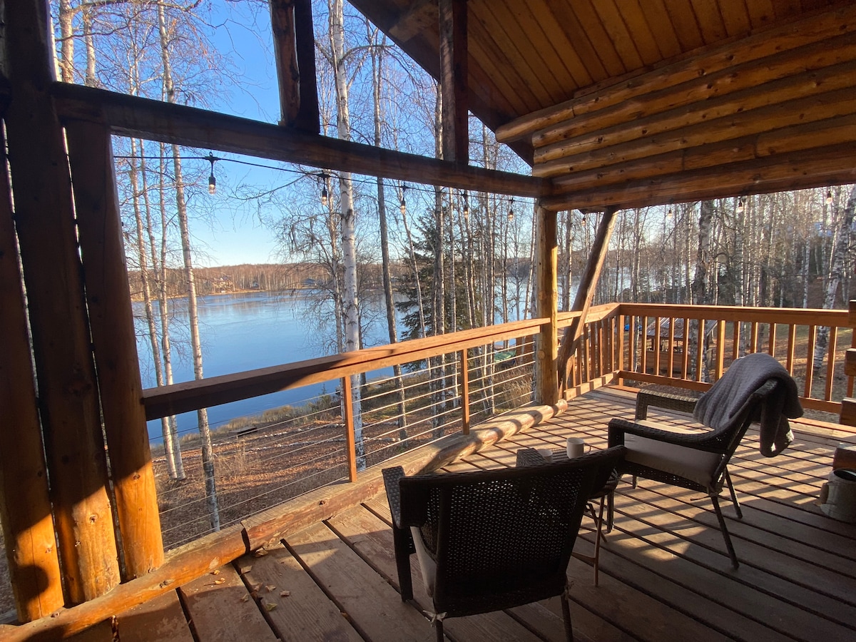 A covered deck space is presented with a view of the serene lake through wooden railings and supports. Two chairs are arranged on the wooden floor, accompanied by a soft blanket draped over one. Sunlight reflects off the calm water, surrounded by birch trees.