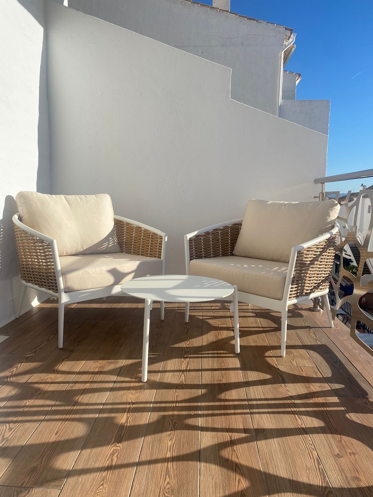 A small outdoor seating area is featured, consisting of two light-colored chairs with woven backrests and comfortable cushions. A minimalistic white table occupies the center, casting shadows on the wooden floor that provides a warm contrast against the bright blue sky.