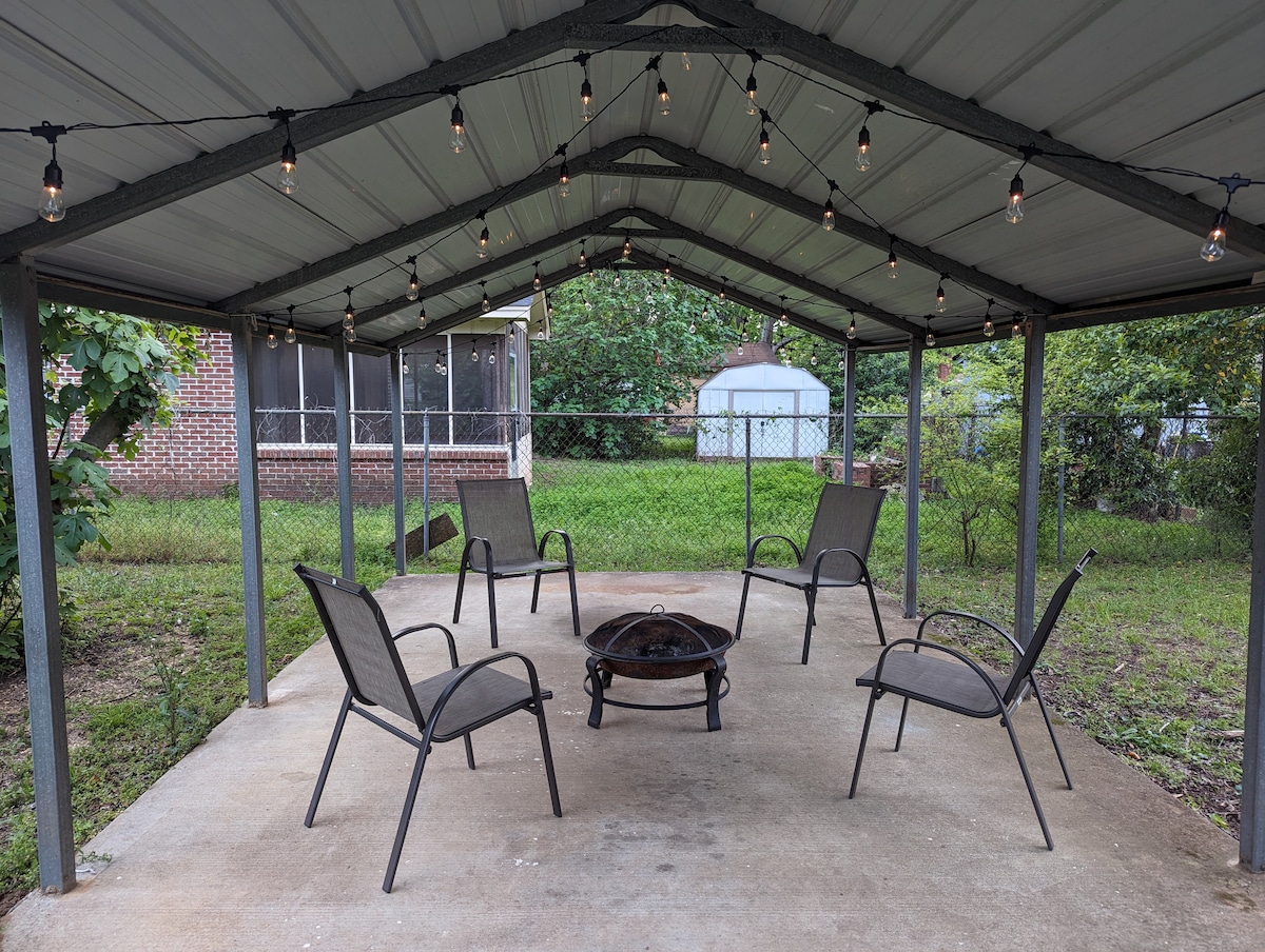 A covered outdoor seating area features four chairs arranged around a central fire pit on a concrete surface. String lights are hung overhead, creating a cozy ambiance for gatherings. A grassy yard and greenery are visible in the backdrop, providing a serene setting.
