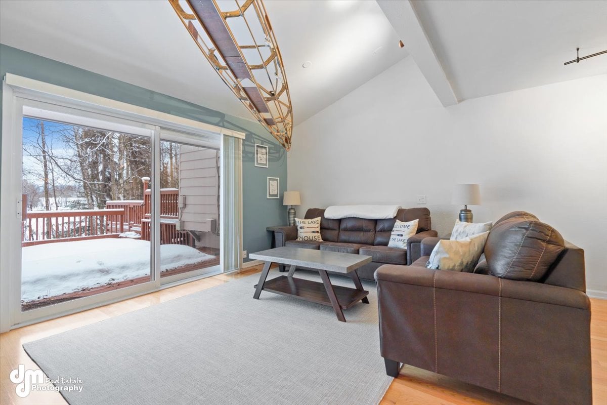 A comfortable living area is presented with a brown leather sofa and accent chairs facing a coffee table. A large sliding glass door allows natural light and reveals a snowy deck outside. A canoe decoratively hangs above the seating area, adding a unique touch.