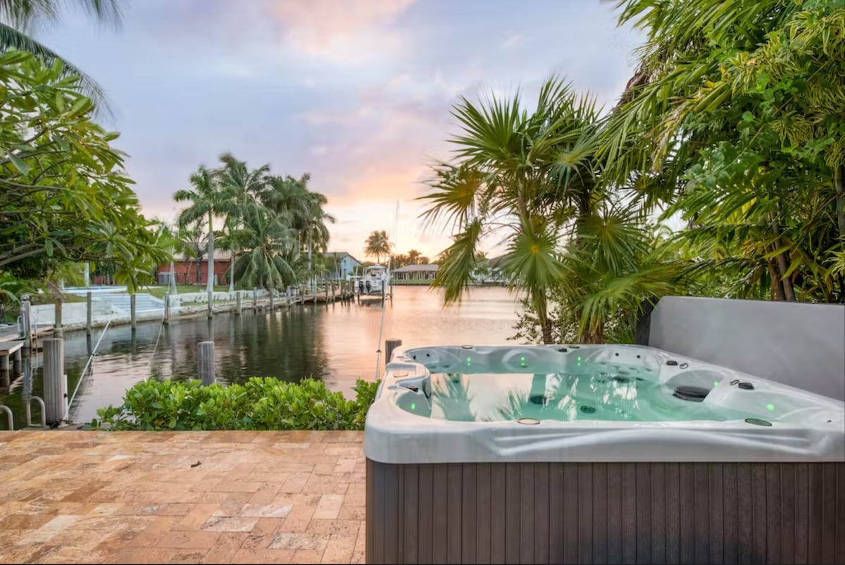 A private hot tub is positioned on a stone patio, overlooking serene water views at sunset. Lush tropical plants frame the scene, enhancing the tranquil ambiance as soft colors of the sky reflect on the water's surface.