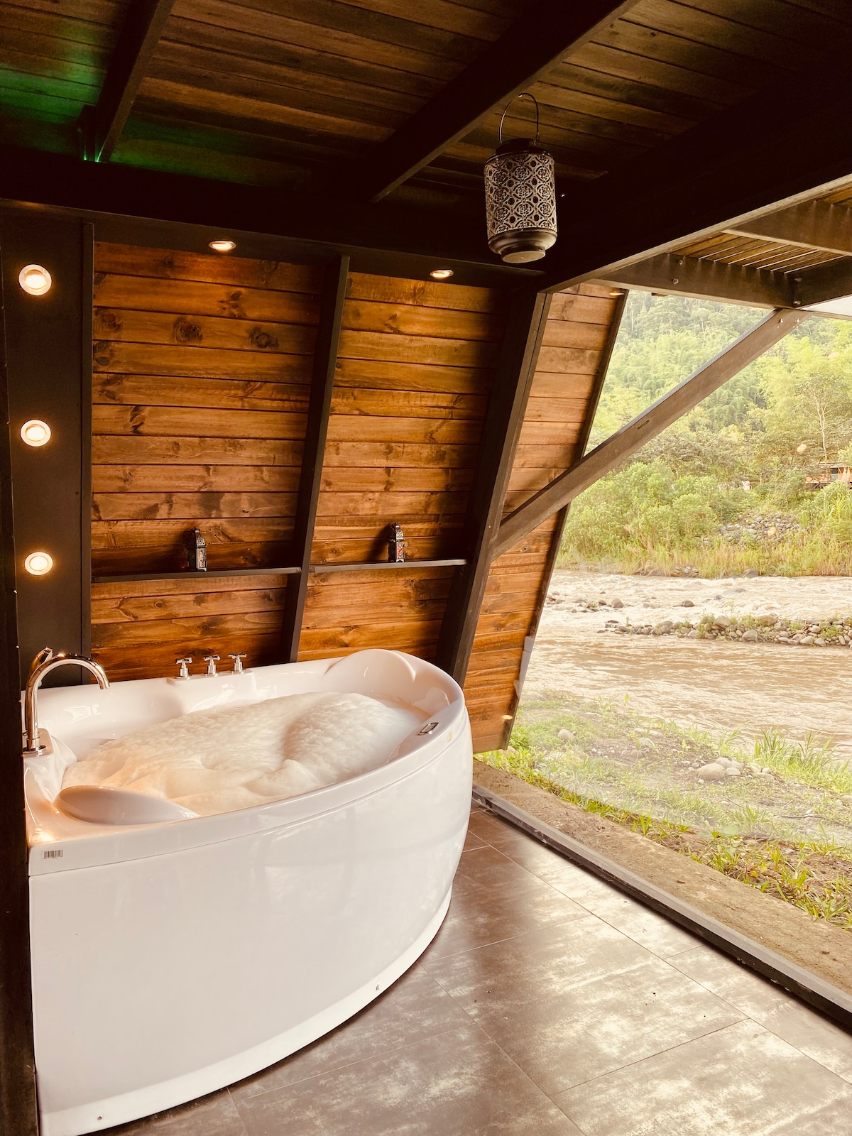 A modern bathroom corner features a freestanding white jacuzzi surrounded by wooden walls. Soft lighting highlights the tub, while a view of the nearby river and lush greenery is visible through the open space.