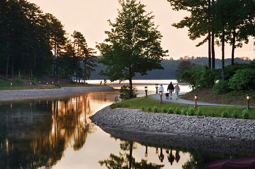A serene lakeside path is lined with trees and shrubs, reflecting tranquil water on either side. Two figures stroll with a dog along the pathway, illuminated by soft lighting. The landscape shows a gradual transition from land to water, with a distant view of the lake.