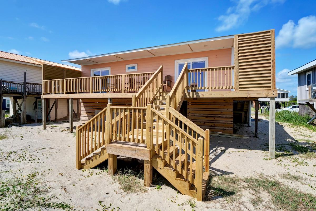 The exterior of a coral-colored cottage is displayed, featuring a spacious wooden deck with multiple steps leading to the entrance. The sandy ground surrounds the cottage, and contrasting light blue skies provide a bright backdrop.