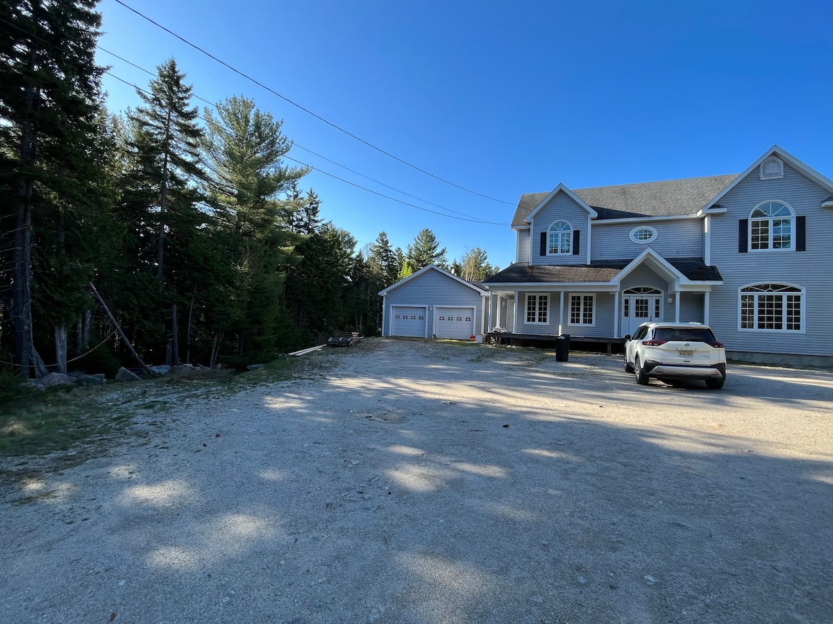 A gravel driveway leads to a large two-story house with multiple windows. The exterior features a light gray color and a well-kept appearance, surrounded by tall trees. A garage is visible to one side, with a white vehicle parked nearby.
