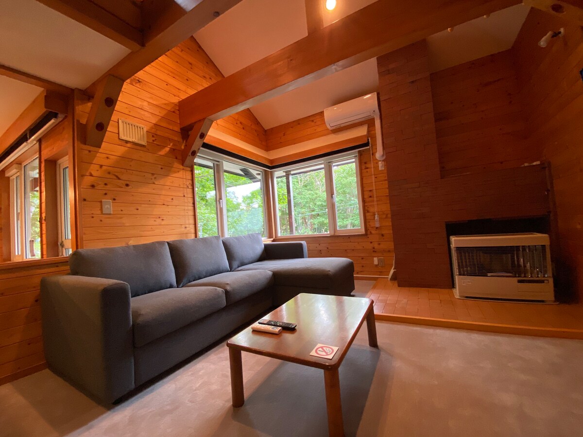 A spacious living area is presented, featuring a comfortable sofa and a wooden coffee table. Large windows allow natural light to fill the room, while a stylish fireplace adds warmth. Wooden walls enhance the cabin's rustic charm, creating a welcoming environment.
