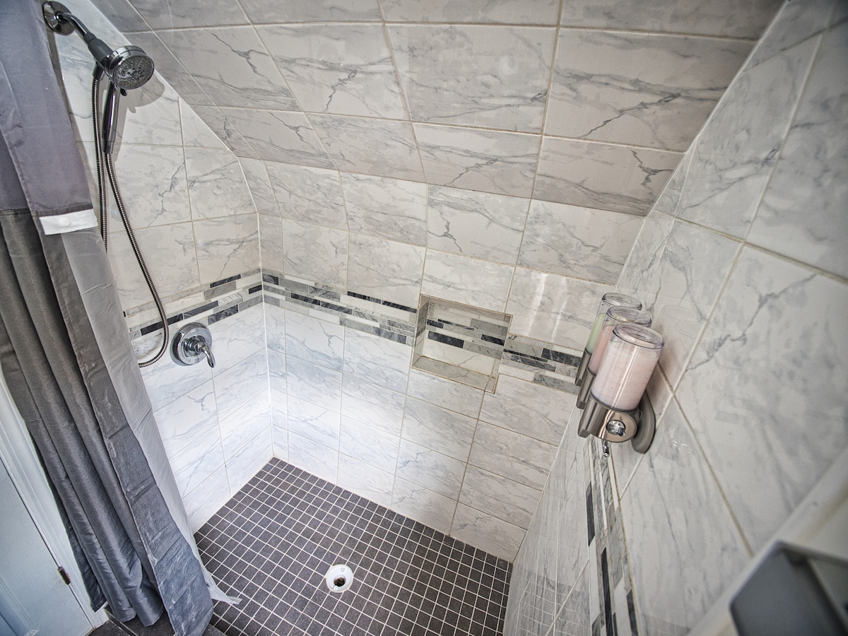 The shower features a sleek design with marble-patterned tile walls and dark tiled flooring. A rainfall showerhead is mounted above, and wall-mounted dispensers are filled with toiletries. The shower's open layout offers ample space for comfortable movement.