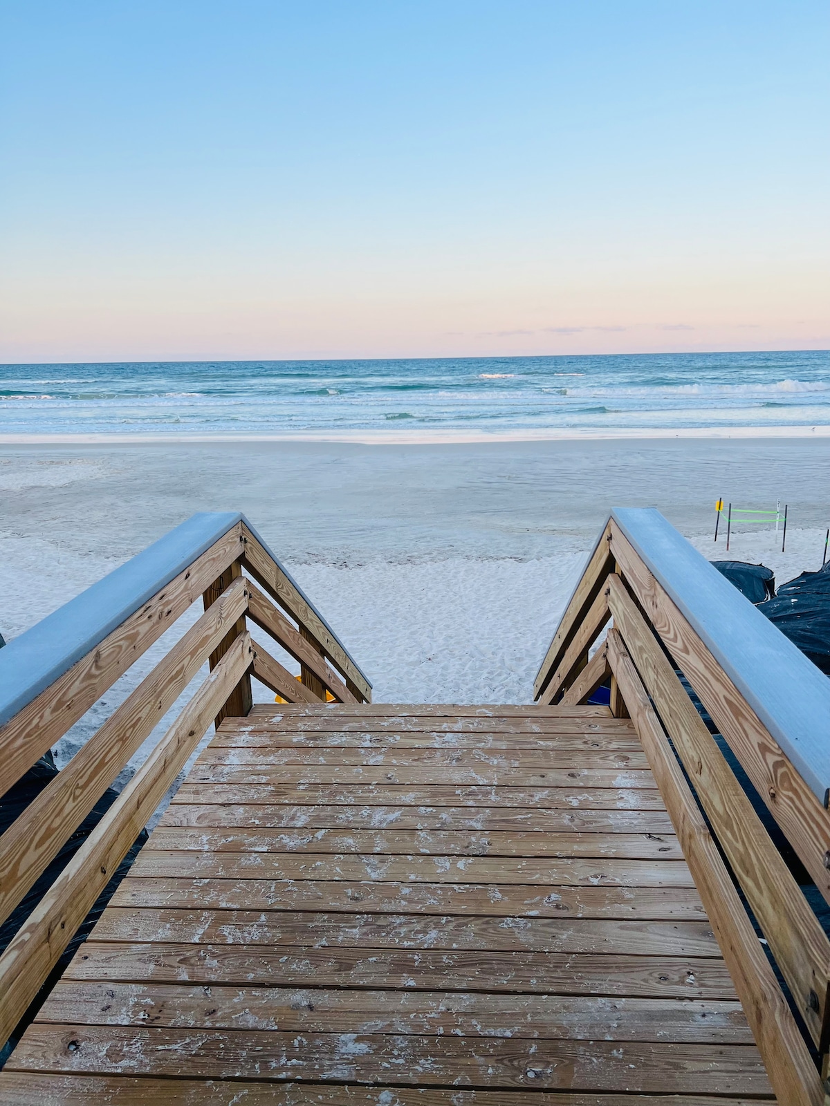 A wooden walkway leads down to the sandy beach, with the ocean visible in the distance. Gentle waves wash against the shore under a pastel sky, transitioning from pink to blue as daylight begins to fade. The scene conveys a sense of tranquility.