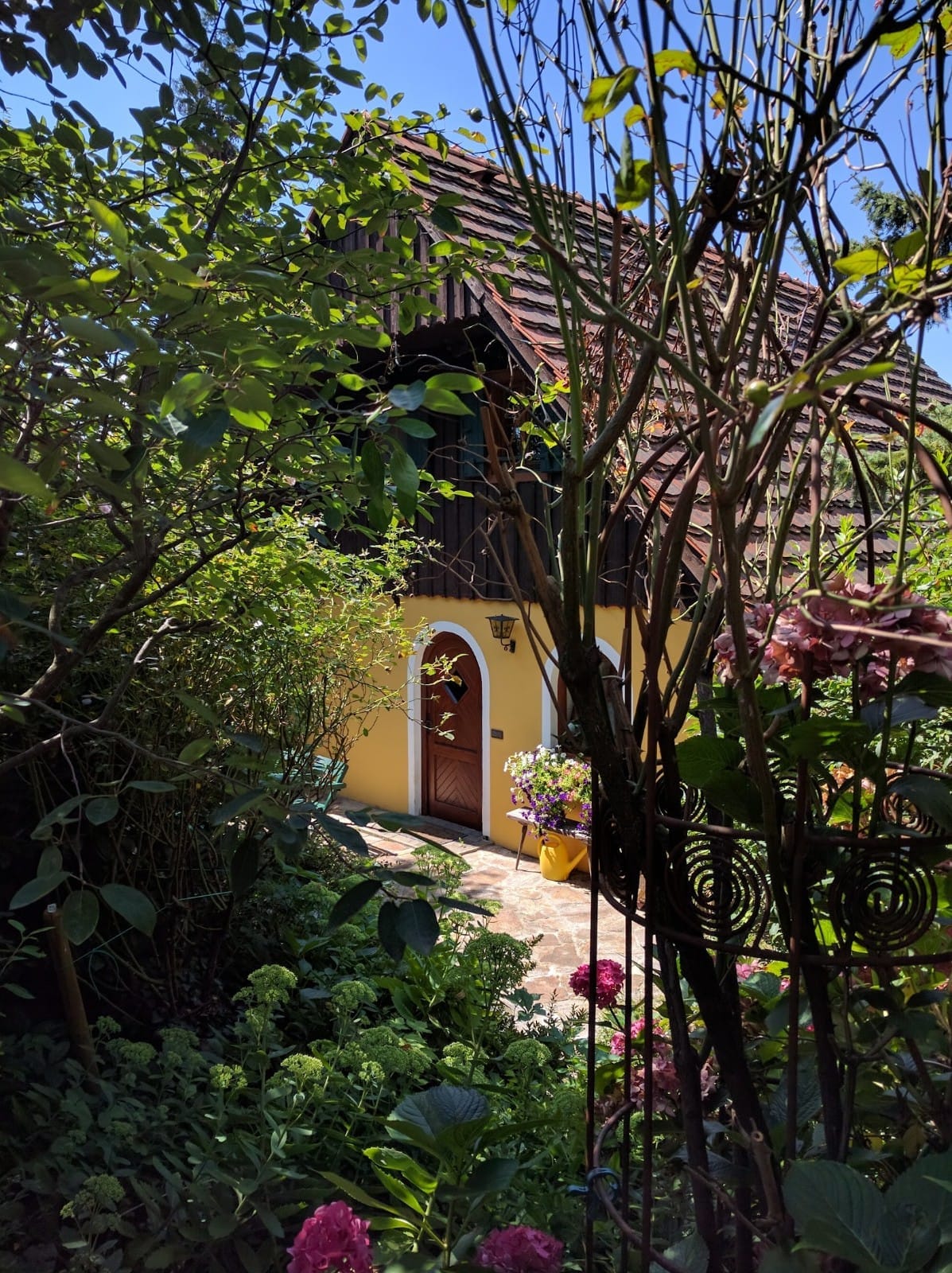 A quaint house is partially obscured by lush greenery and colorful flowers. The entrance features a rounded door framed by decorative elements and surrounded by vibrant plants, inviting a sense of calm and tranquility.