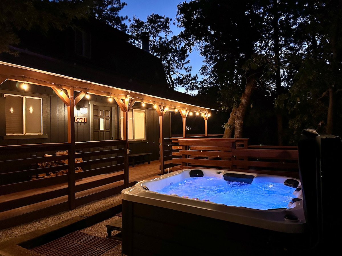 A hot tub is illuminated with soft blue light on the deck of a cabin. Surrounding trees provide privacy, while string lights along the deck create a warm ambiance. The cabin features large windows, suggesting an inviting indoor space.