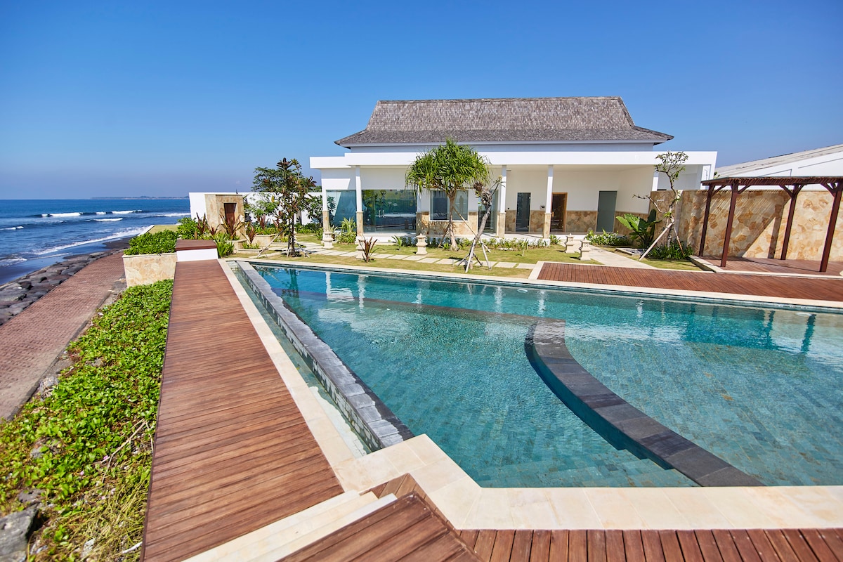 A modern villa is set against the ocean, featuring a glass-walled infinity pool that appears to extend into the horizon. Surrounding the pool, a wooden deck leads through landscaped gardens, providing a serene outdoor space. The clear blue sky complements the tranquil coastal scene.