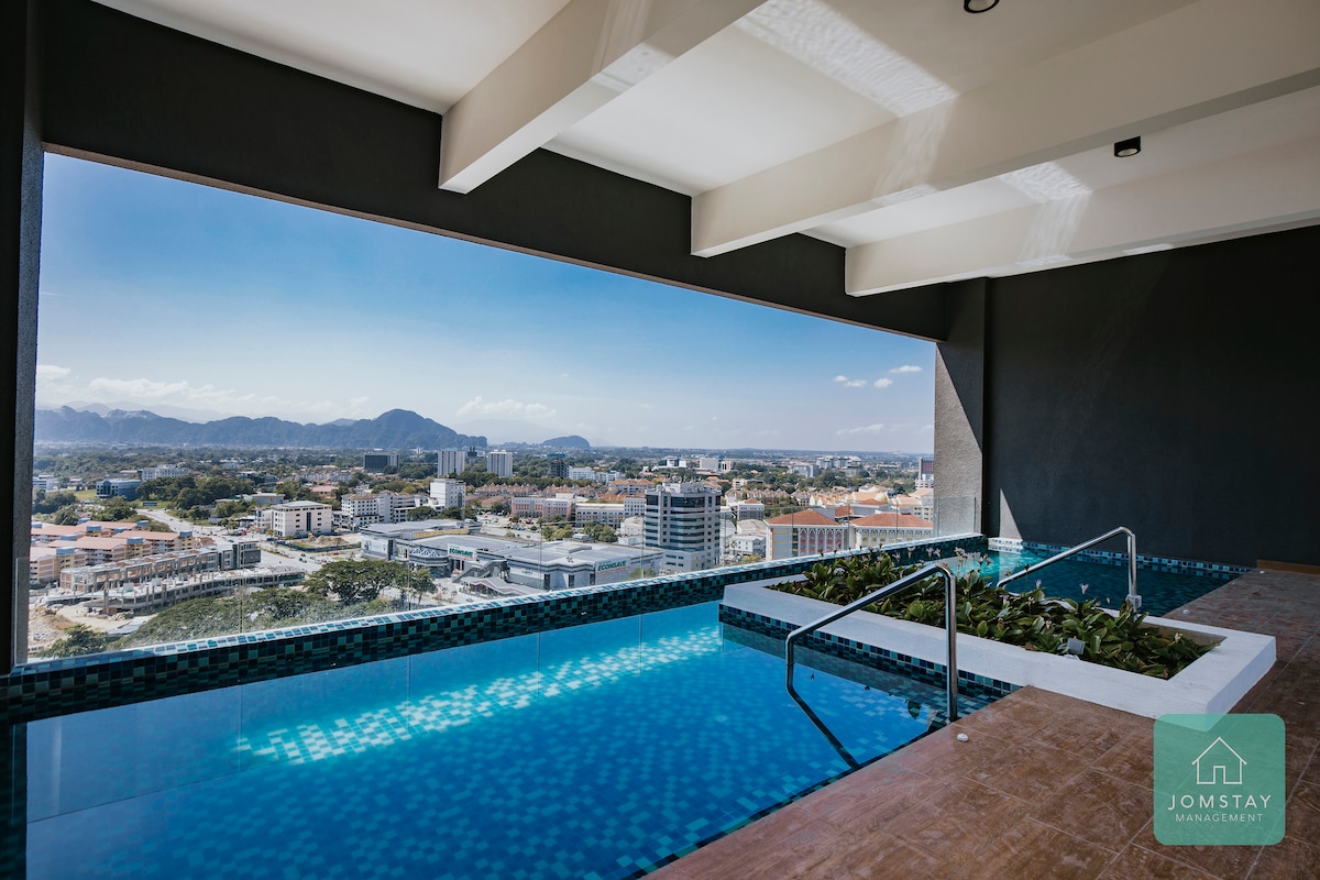 An outdoor swimming pool is showcased, featuring clear blue water and a panoramic view of the surrounding cityscape and mountains. Green landscaping is visible along the edge of the pool, while the pool area is illuminated by natural light from a large open space above.
