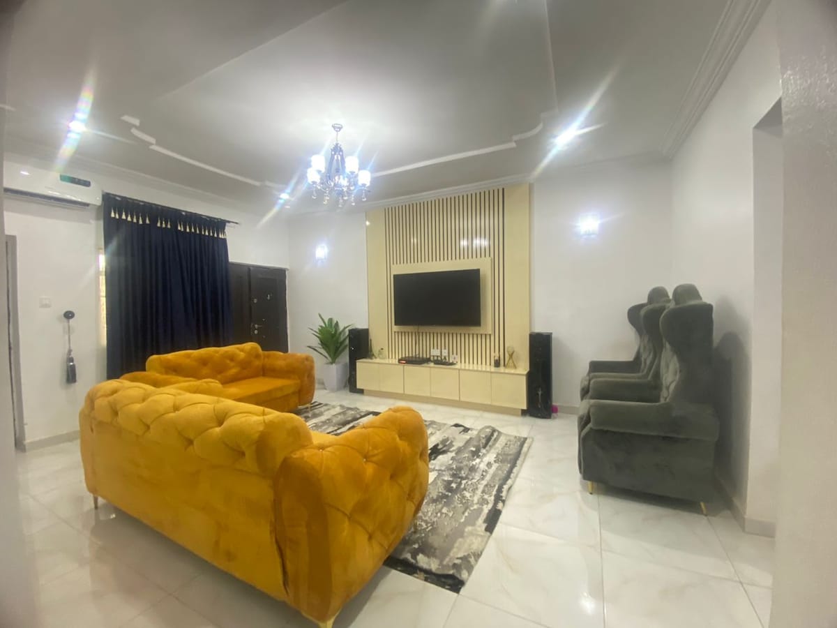 A spacious living room is presented with plush, yellow and dark green sofas arranged around a decorative rug. A flat-screen television is mounted on a stylish wall unit. A large window is dressed with dark curtains, and a potted plant adds a touch of greenery.