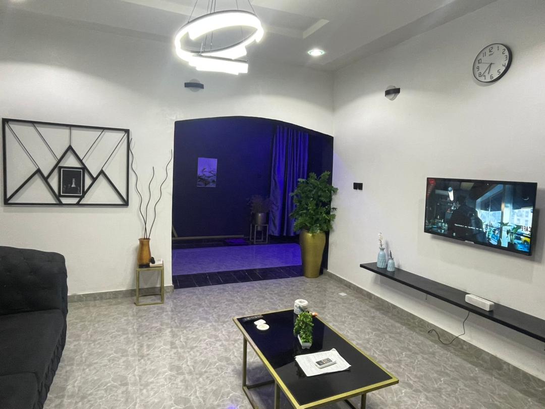 A modern living area features a dark sofa, a coffee table with a small plant, and a wall-mounted television. A decorative clock hangs above, while an entrance leads to a dimly lit space with curtains and a plant in a decorative pot.