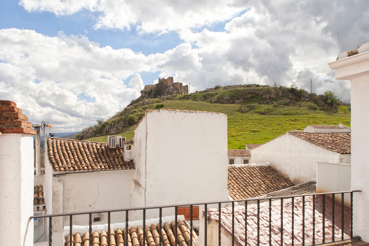 A view from a balcony features whitewashed buildings with terracotta roofs leading toward a hillside. The medieval castle is visible atop the hill, surrounded by greenery under a partly cloudy sky.