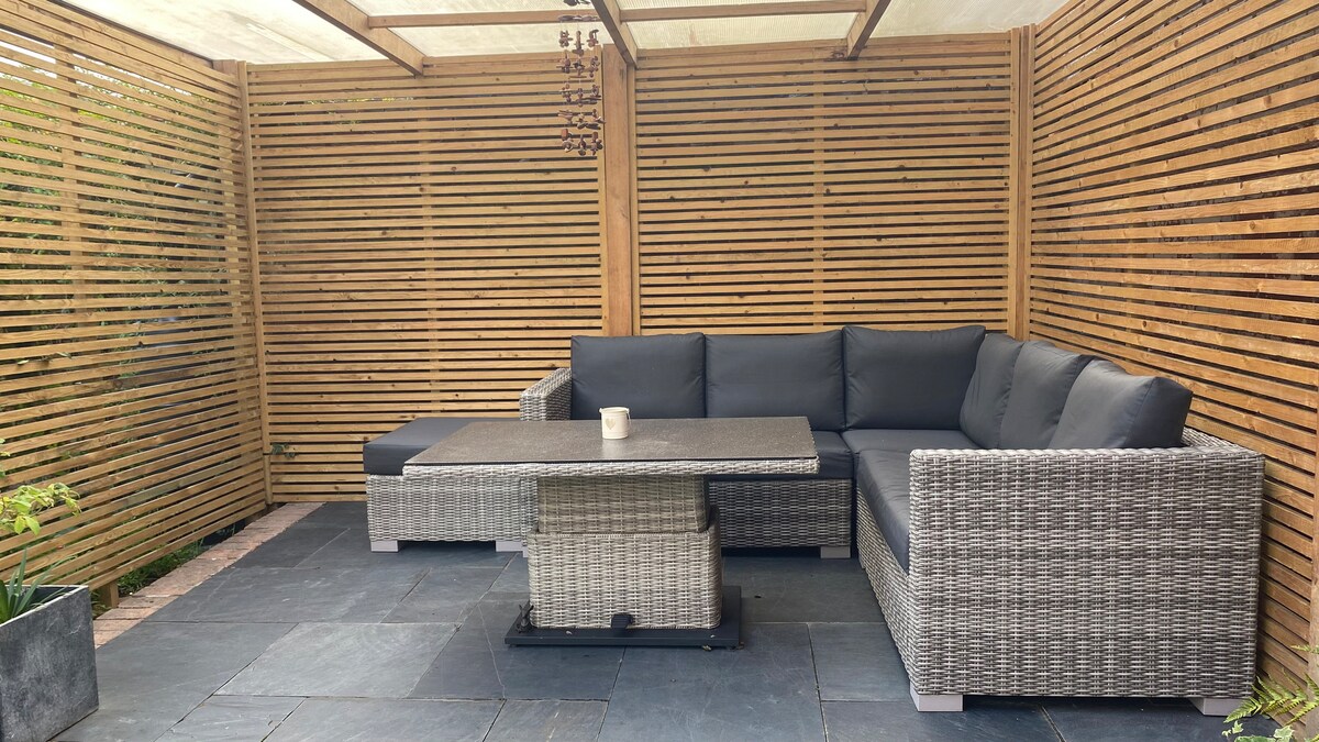 A covered outdoor seating area features a spacious, sectional sofa with dark cushions surrounding a low table. The wood slat walls provide privacy, while a grey stone floor adds a contemporary touch to this inviting space.
