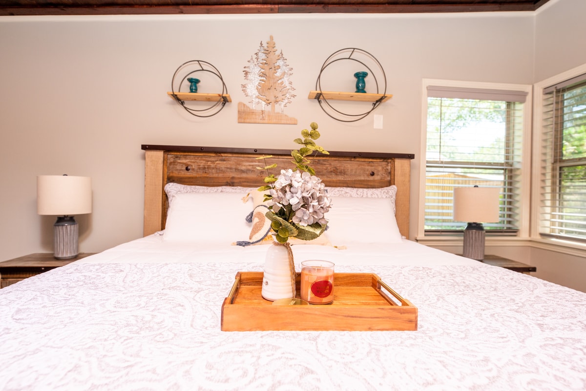 A spacious bed is centered against a wooden headboard, with light bedding and decorative pillows. Soft lamps flank the bed on each side, and a wooden tray holds a drink and floral arrangement at the foot. Natural light streams through windows with blinds.