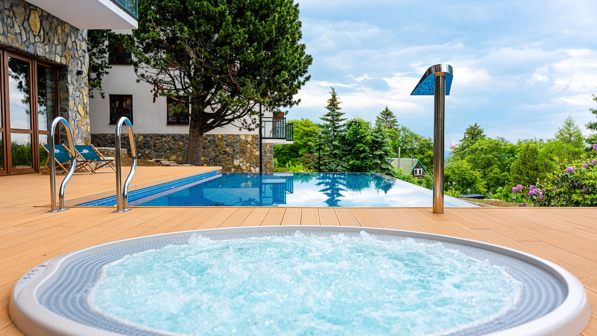 A serene outdoor area features a heated jacuzzi with bubbling water, situated beside an inviting pool. Lush greenery surrounds the space, enhancing the natural setting. Lounge chairs are positioned nearby, offering spots for relaxation under the sky.