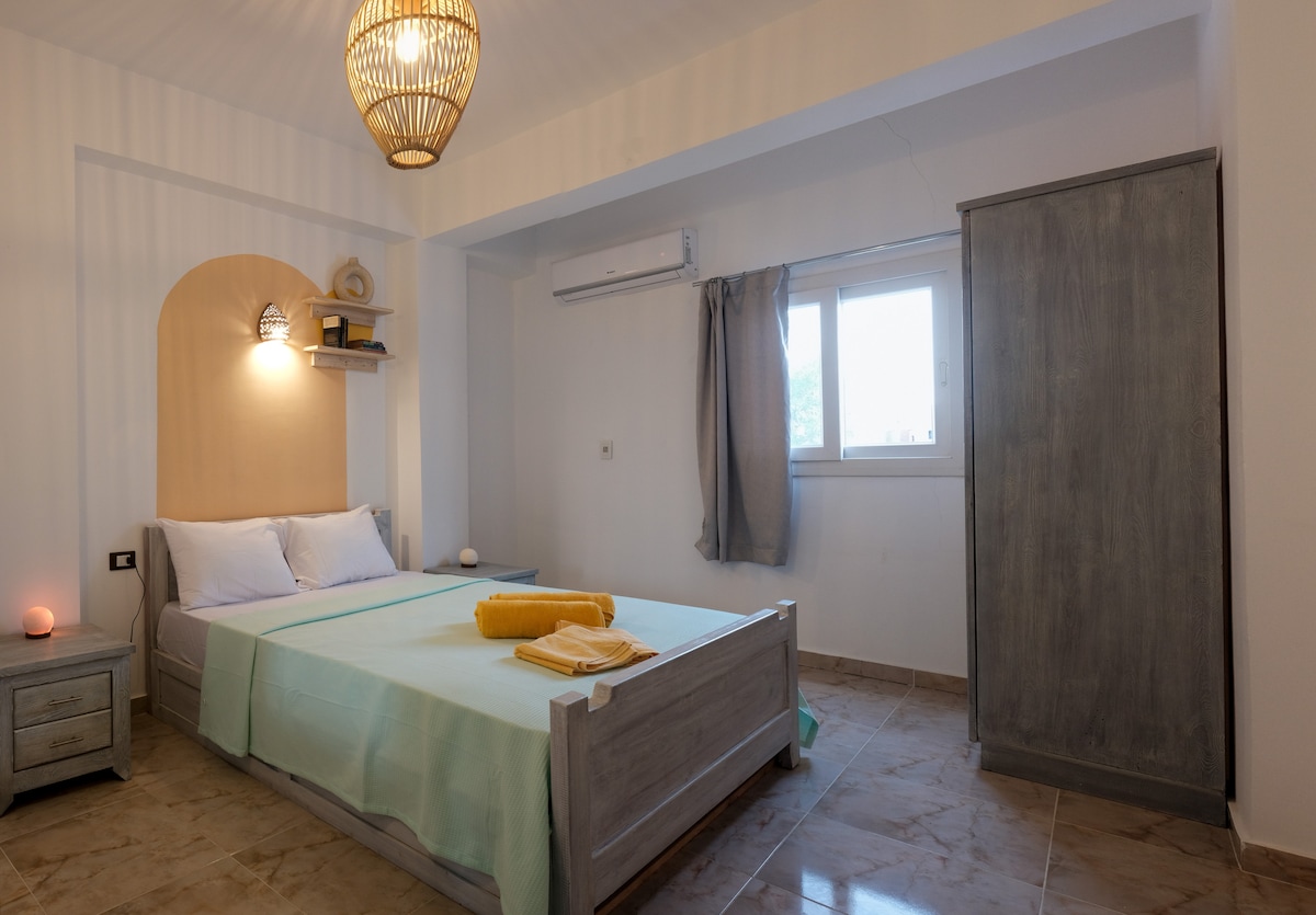 A well-lit sleeping area features a spacious bed adorned with light blankets. A large window with sheer curtains allows natural light to enter. Neutral tones create a calming ambiance, complemented by a wooden wardrobe and a decorative light fixture above.