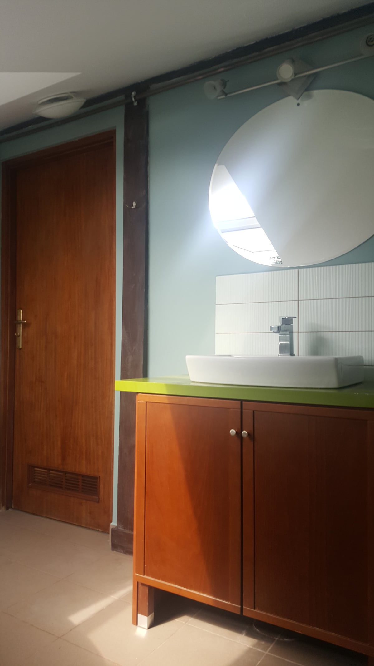 A modern bathroom is displayed with a circular mirror above a white sink, set against a light blue wall. A wooden cabinet with two doors provides storage beneath the sink. A wooden door leading elsewhere is visible, adding to the space's functional design.