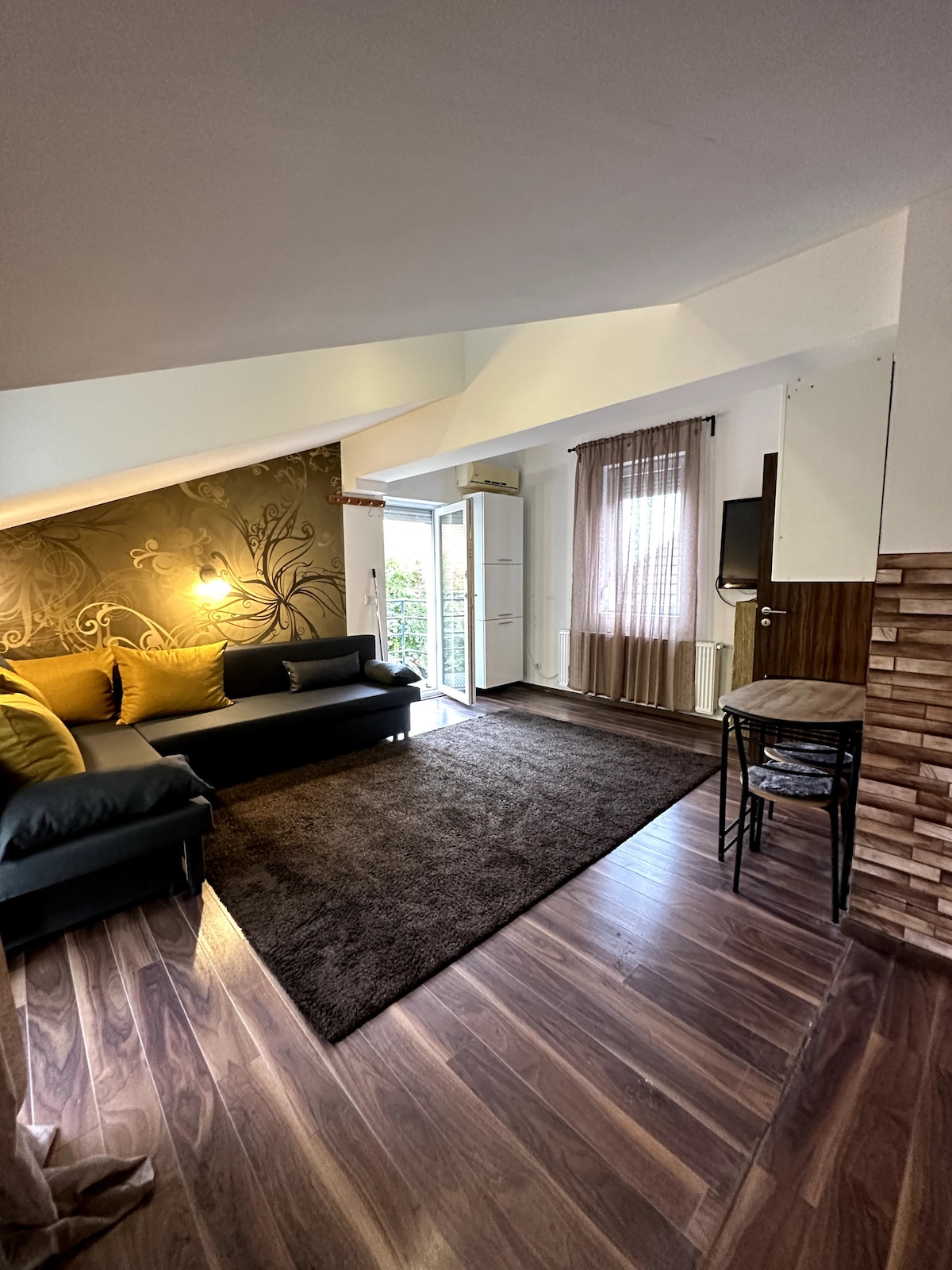 A comfortably arranged living area features a dark area rug over hardwood flooring. A black sofa adorned with yellow cushions invites relaxation. Natural light enters through the glass door leading to an outdoor space, while a flat-screen television is mounted on the wall.
