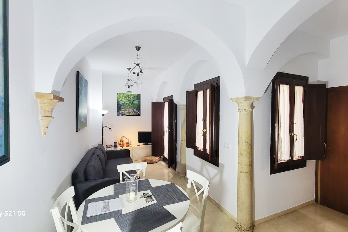 A spacious living area features arched ceilings and classic columns, with natural light streaming through the windows. A comfortable sofa is positioned near a television, while a dining table with white chairs is set up for meals. Neutral tones create a calming environment.