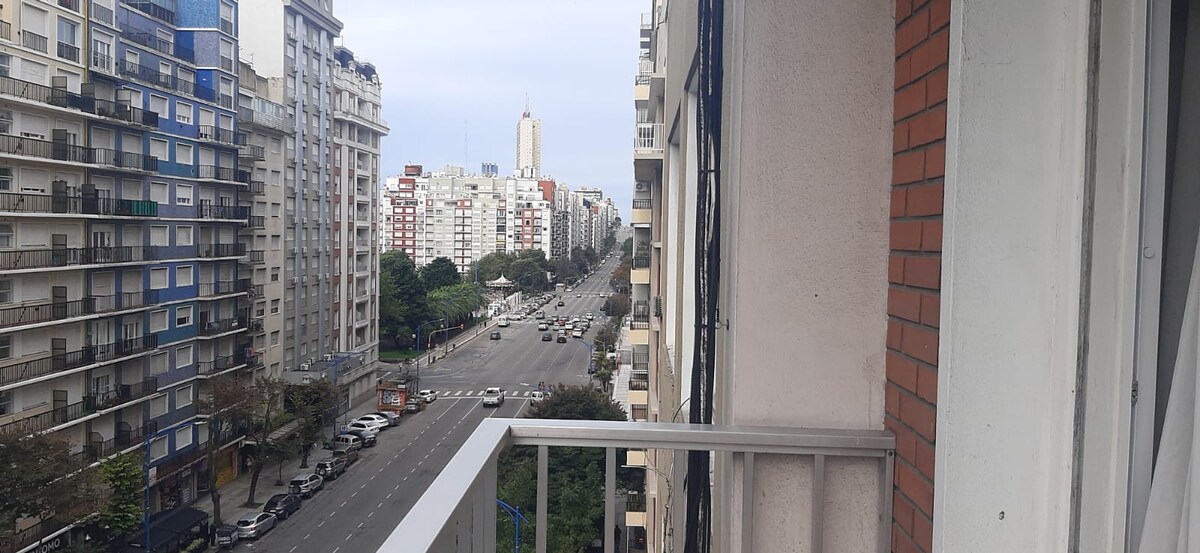 A balcony view showcases a bustling city street lined with tall buildings. Traffic can be seen moving along the street, with green trees dotting the landscape. In the distance, a tall tower rises above the surrounding architecture.