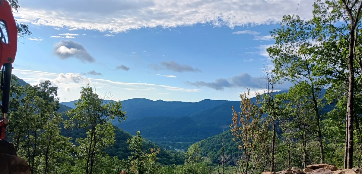 The image captures a wide view of rolling green mountains under a blue sky, interspersed with fluffy white clouds. Lush trees frame the foreground, highlighting the expansive, natural landscape that surrounds the area.