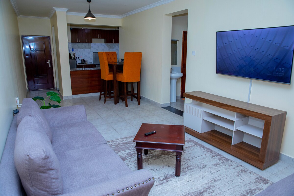 A modern living area is showcased with a comfortable grey sofa and a wooden coffee table. A flat-screen TV is mounted on a light wood media console. The open-plan space flows into a kitchen area featuring tall chairs around a counter, with light-colored cabinets in the background.