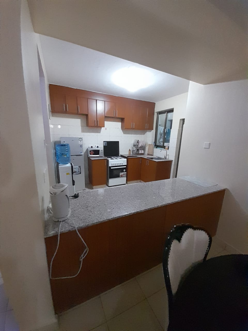 A spacious kitchen area is presented, featuring wooden cabinets and a gray granite countertop. Essential appliances are visible, including a stove and refrigerator. A water dispenser is positioned on the counter, with ample natural light filtering through the nearby window.