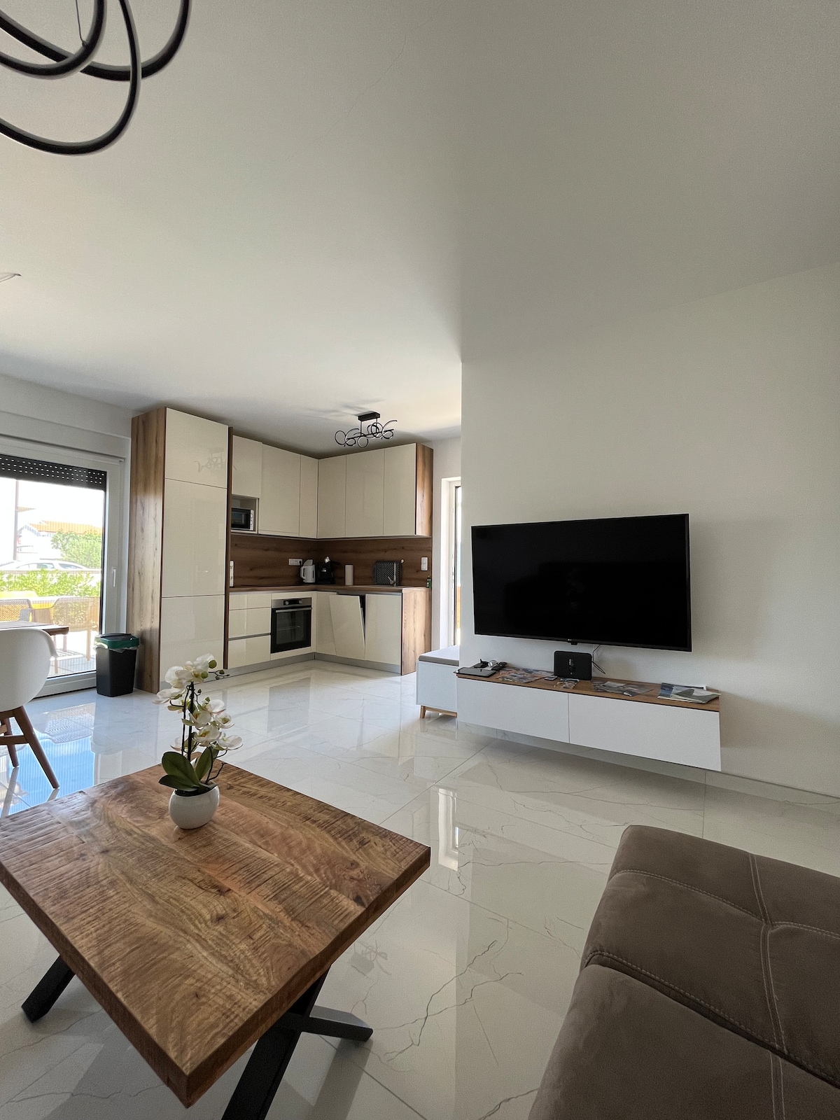 A modern living space is showcased with light-colored walls and glossy tiled floors. A stylish wooden coffee table is positioned in front of a comfortable sofa. The television is mounted on a white cabinet, and a well-equipped kitchen area is visible in the background.