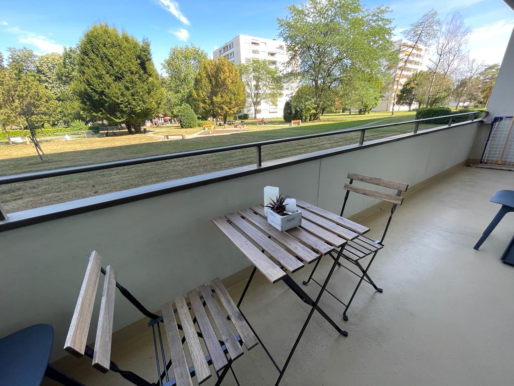 A small balcony is furnished with a wooden table and two folding chairs. A vase with a plant is positioned on the table. The balcony overlooks a grassy playground area with trees and low buildings in the background, providing a peaceful outdoor view.