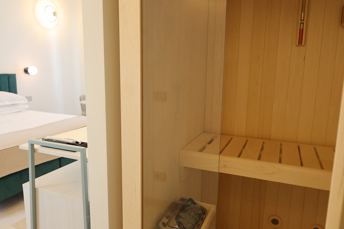 A sauna is showcased within a glass enclosure, featuring wooden slats and a thermometer. The inviting warmth of the sauna contrasts with the light-colored walls and furnishings visible in the adjacent room.