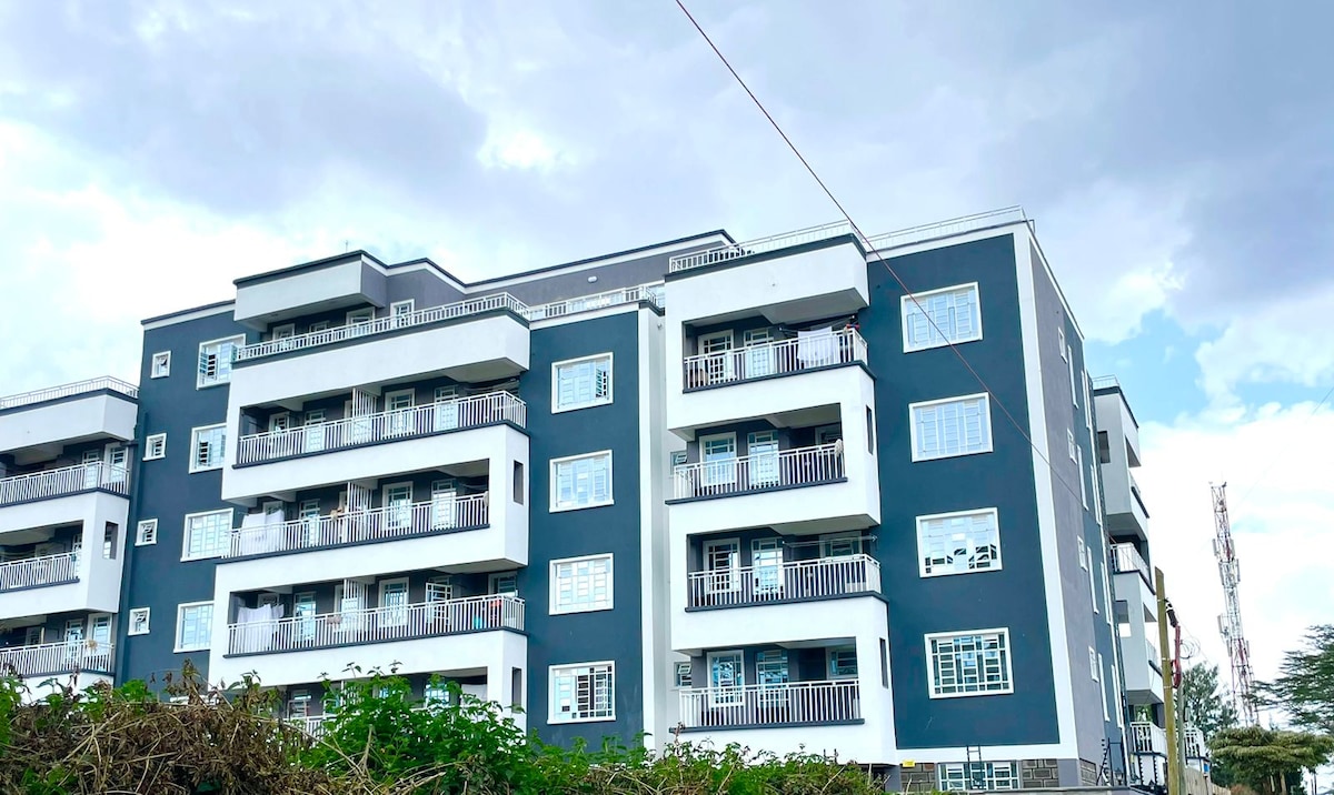 The exterior of a modern apartment building is displayed, featuring a mix of dark blue and white facades. Multiple balconies with white railings line the structure, and large windows allow natural light to enter the units. Surrounding greenery adds a natural touch.