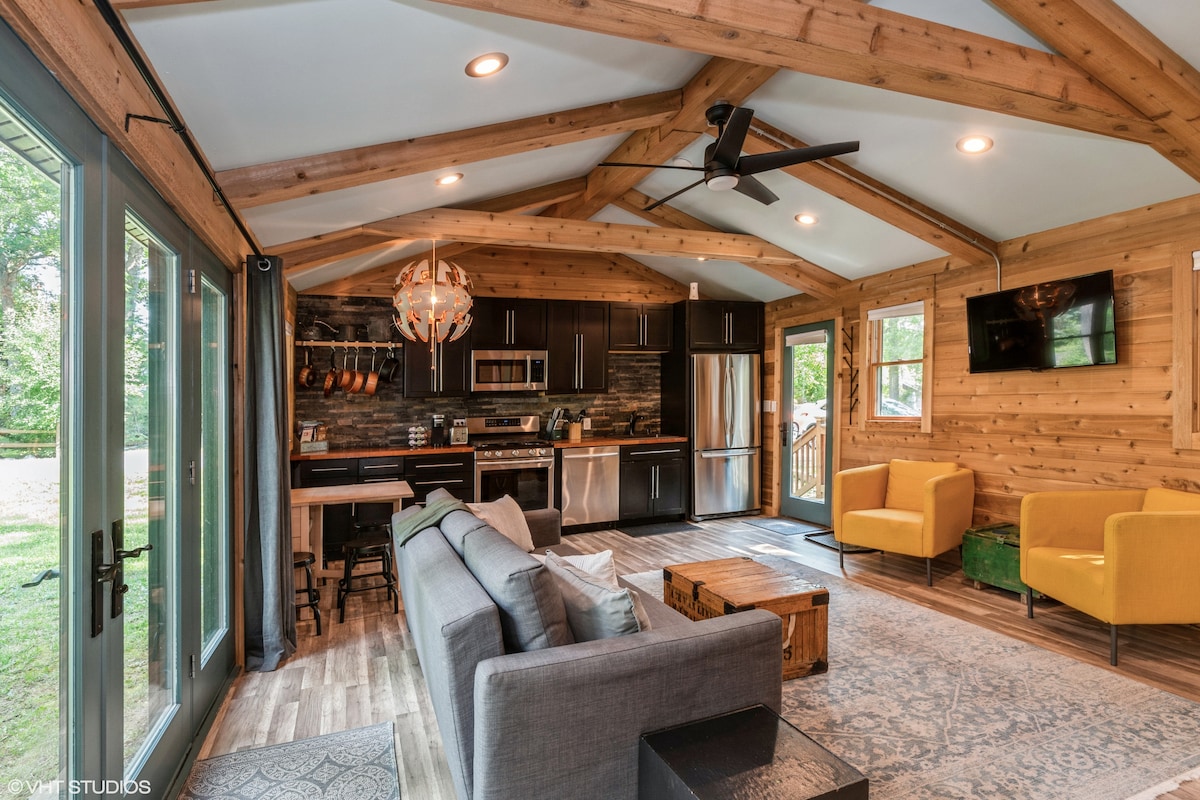 The living area features a cozy gray sofa facing a wooden coffee table, paired with two bright yellow armchairs. Exposed wooden beams create a spacious feel under the vaulted ceiling. A well-equipped kitchen with modern appliances is visible in the background, complemented by a dining area.