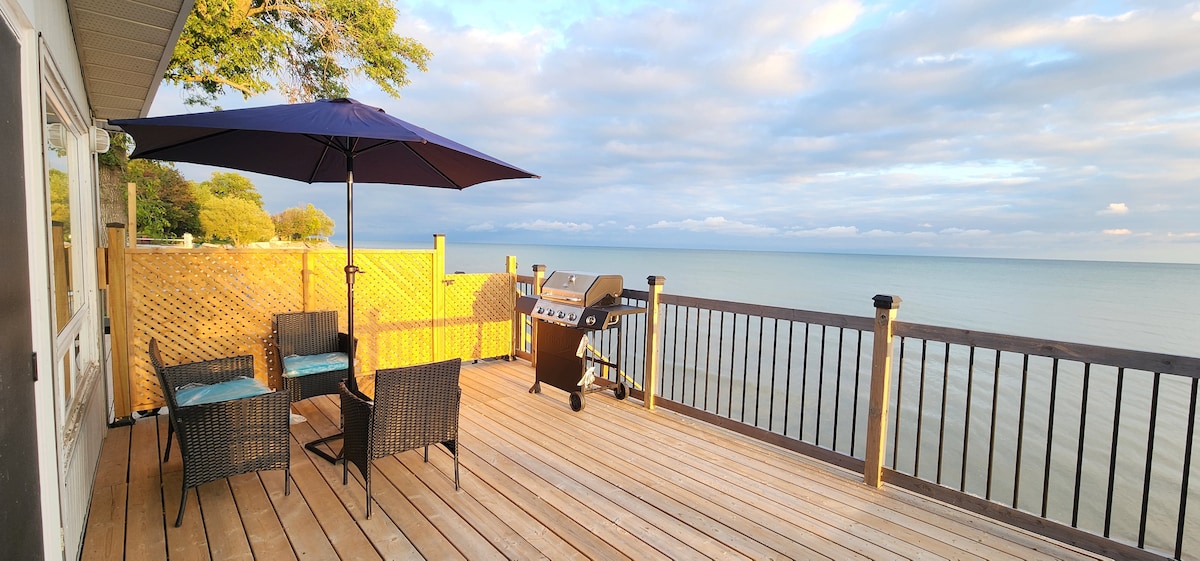 A spacious deck overlooks serene waters, featuring a patio table with two chairs and a large umbrella providing shade. A grill is positioned nearby, offering an ideal space for outdoor enjoyment. The calm lake reflects the sky, enhancing the tranquil setting.
