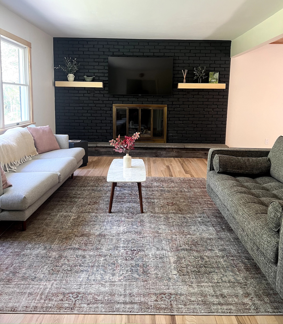 A living area features a modern gray sofa and a light-colored loveseat. A coffee table with a simple vase is centered on an area rug. A black brick fireplace adds character, while a mounted TV and decorative shelves provide an inviting setting.