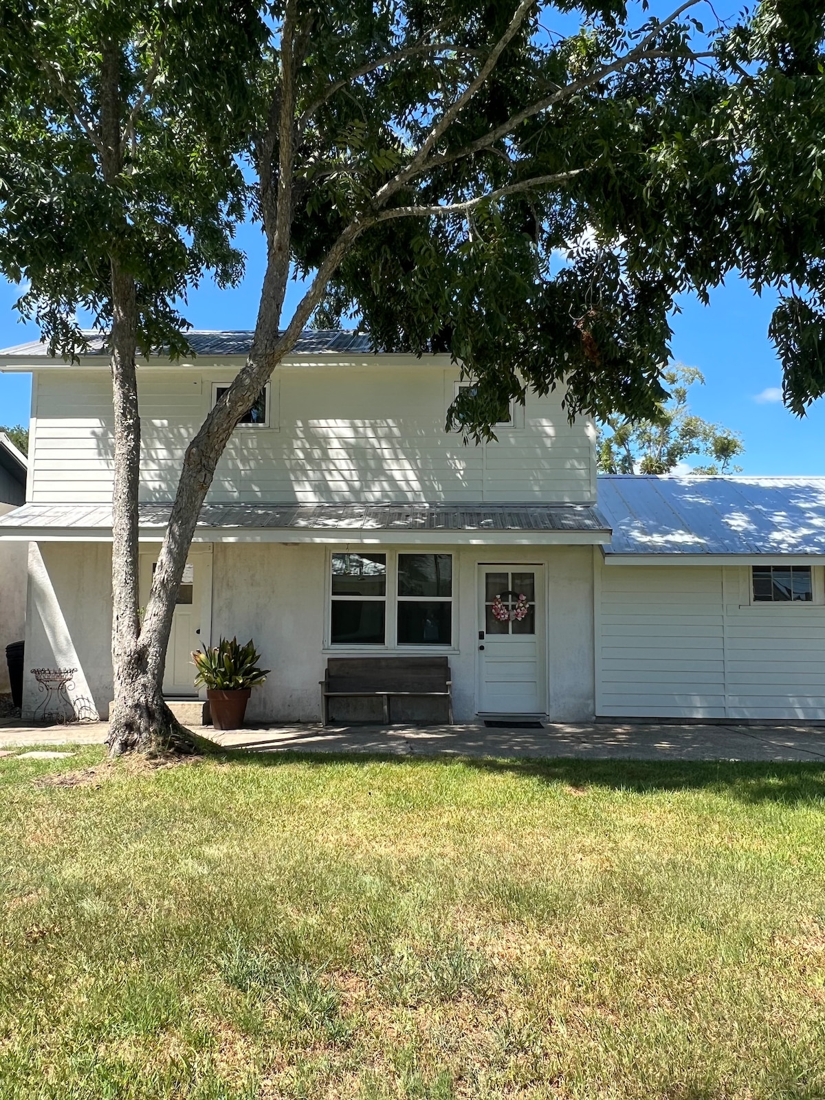 The exterior of a two-story unit is visible, featuring a white facade and a metal roof. A large tree provides shade in the front yard, complemented by a potted plant near the entrance. A garage door is situated on the right side.