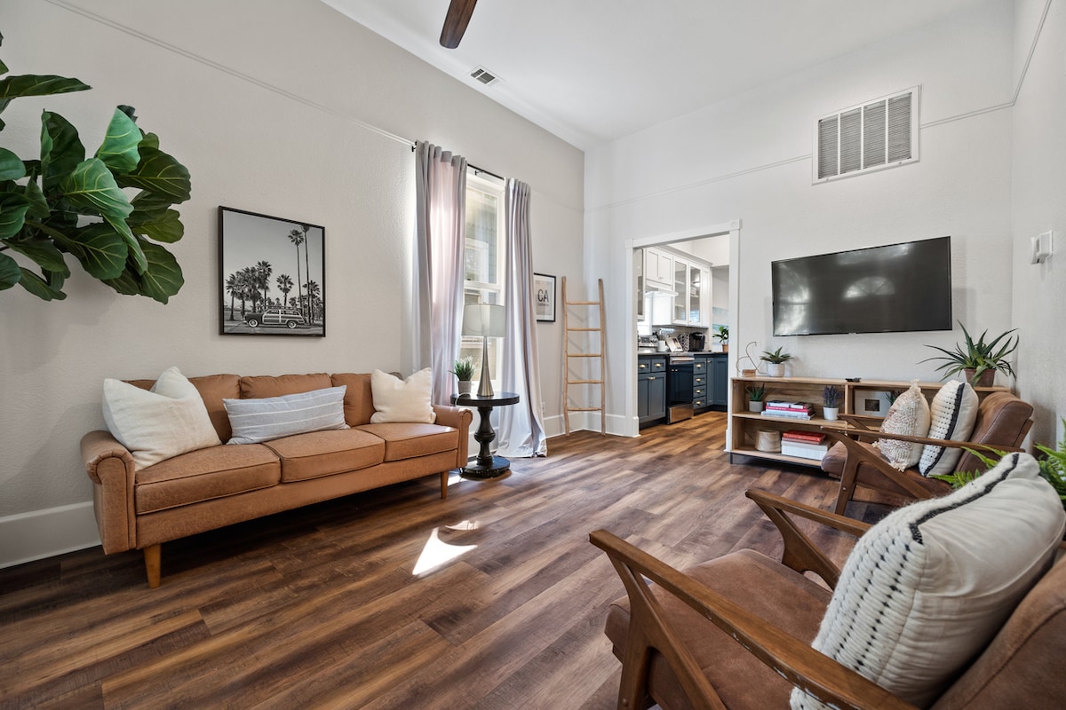 A warm and inviting living area features a comfortable brown sofa, two wooden chairs, and a small side table. Natural light floods the space through large windows, with soft curtains gently framing them. A large-screen TV is mounted on the wall, and decorative plants add a touch of greenery.