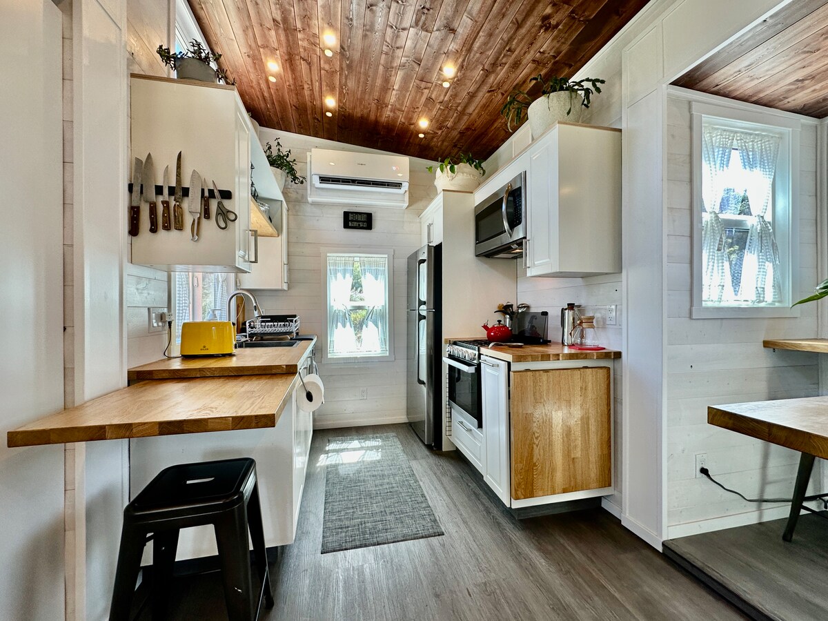 A compact kitchen features modern appliances including a refrigerator, stove, and microwave. Natural light enters through two windows adorned with sheer curtains. Wooden accents are present on the countertops and dining area, with kitchenware neatly organized on display. A small table is set for dining.