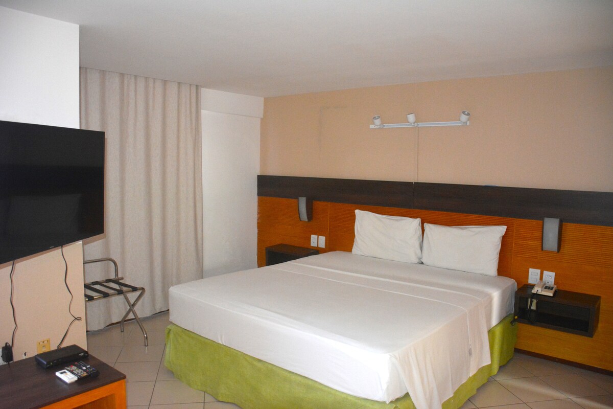 A spacious sleeping area is equipped with a large bed featuring crisp white linens. A flat-screen TV is mounted to the wall across from the bed. Natural light filters through light-colored curtains, creating a calm ambiance in the room.