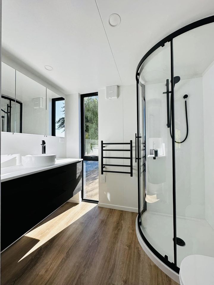 A modern bathroom features a spacious shower enclosed in a curved glass design. The sleek, dark cabinet provides storage, while a large mirror enhances the bright ambiance. Natural light enters through a tall window, illuminating the neutral color scheme and wooden flooring.