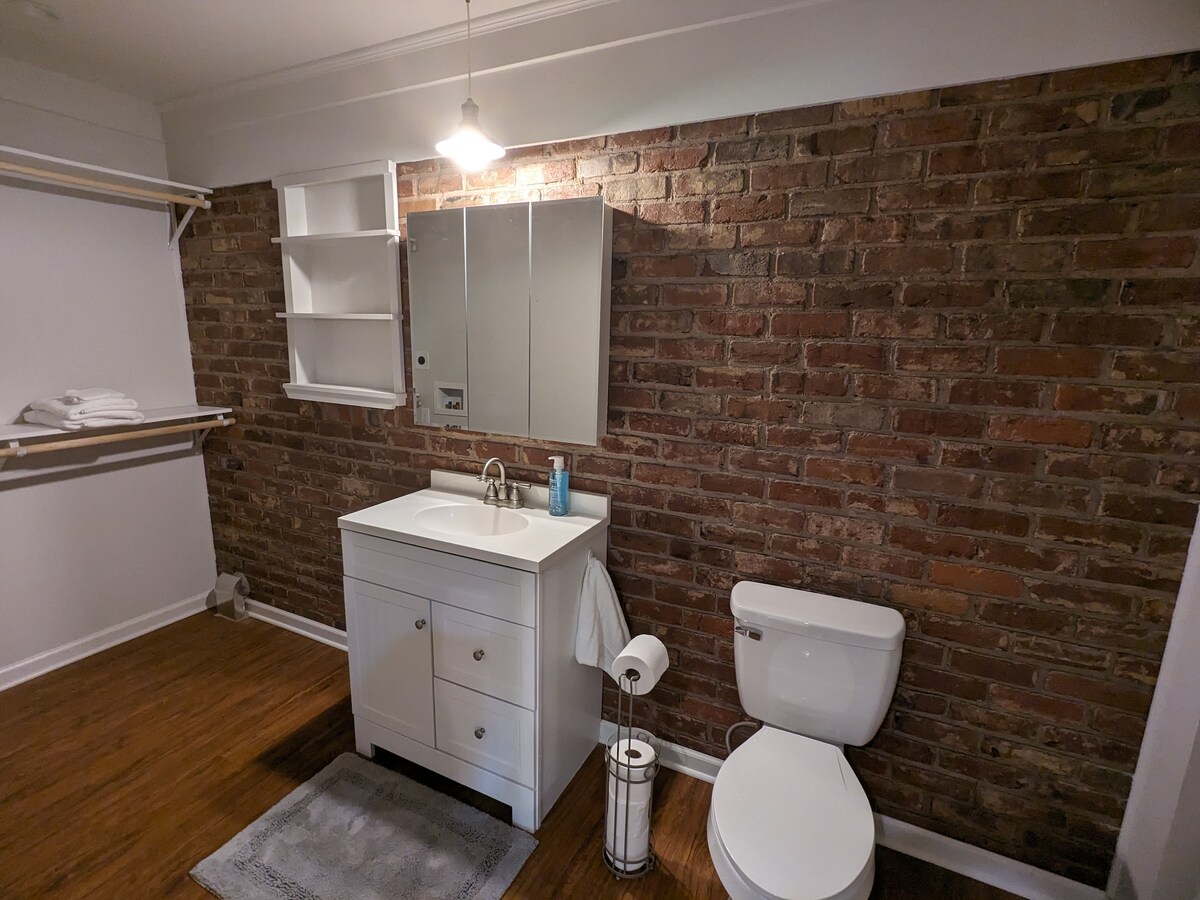 A modern bathroom features a white vanity with a rectangular sink and storage beneath. A large mirror above reflects the brick wall, which adds texture to the space. A white toilet is positioned beside the vanity, and shelves are available for additional storage.