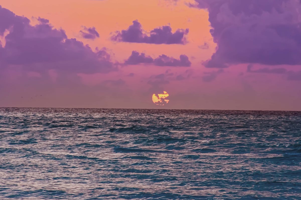 A vibrant sunset casts hues of purple and orange across the sky, reflected on the tranquil waters of the ocean. Waves gently lap at the shore, and the sun appears to be setting on the horizon, creating a serene and picturesque scene.