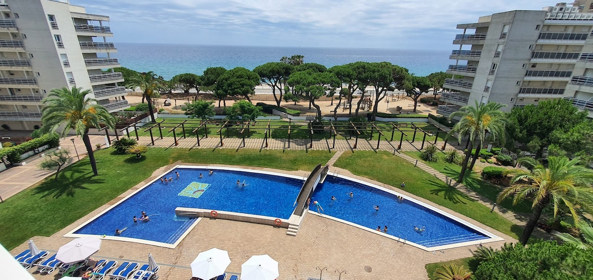 The image captures a large communal swimming pool surrounded by palm trees and lounge chairs. The pool is shaped irregularly, with swimmers enjoying the water. In the background, a sandy beach and the ocean are visible, framed by neatly manicured greenery.