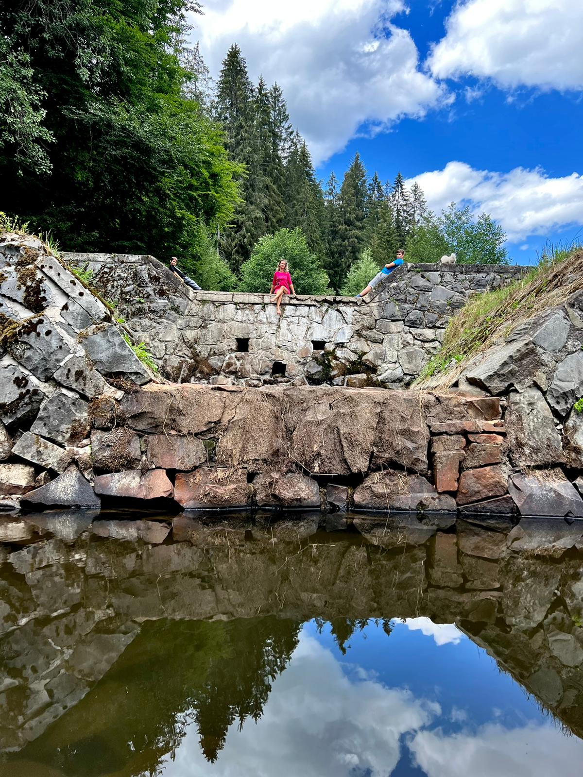 A tranquil pond reflects a stone structure surrounded by lush greenery and towering trees under a partly cloudy sky. Two individuals can be seen at the edge of the stone formation, adding a sense of scale to the natural setting.